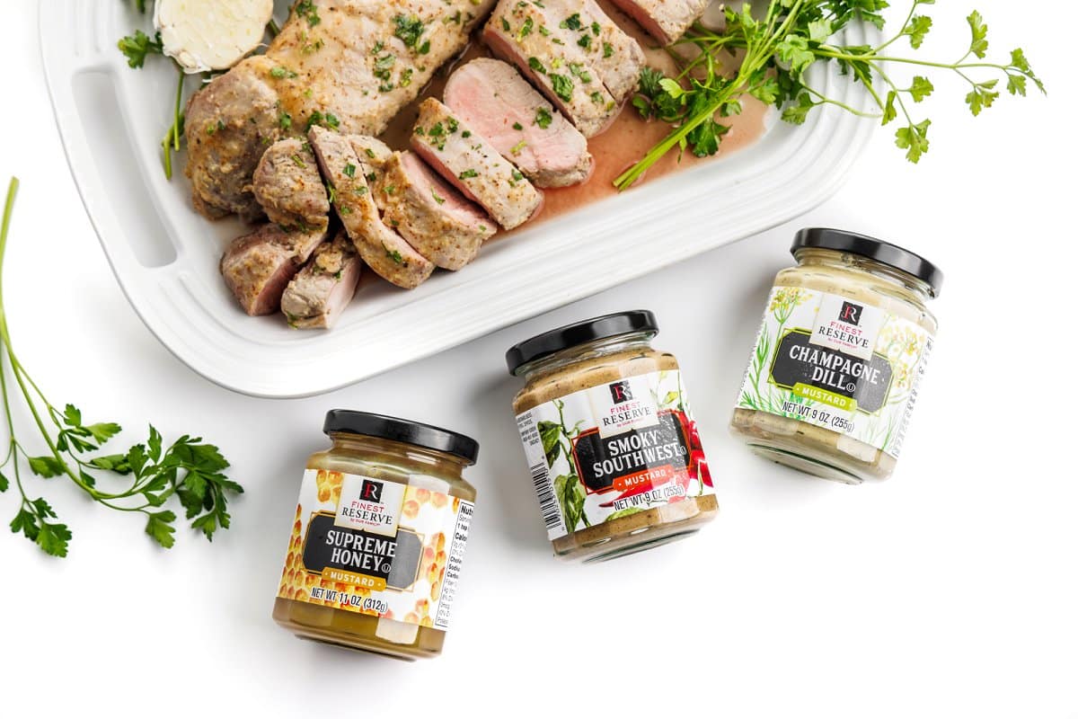 Cooked pork tenderloin on a platter next to containers of Finest Reserve mustard.