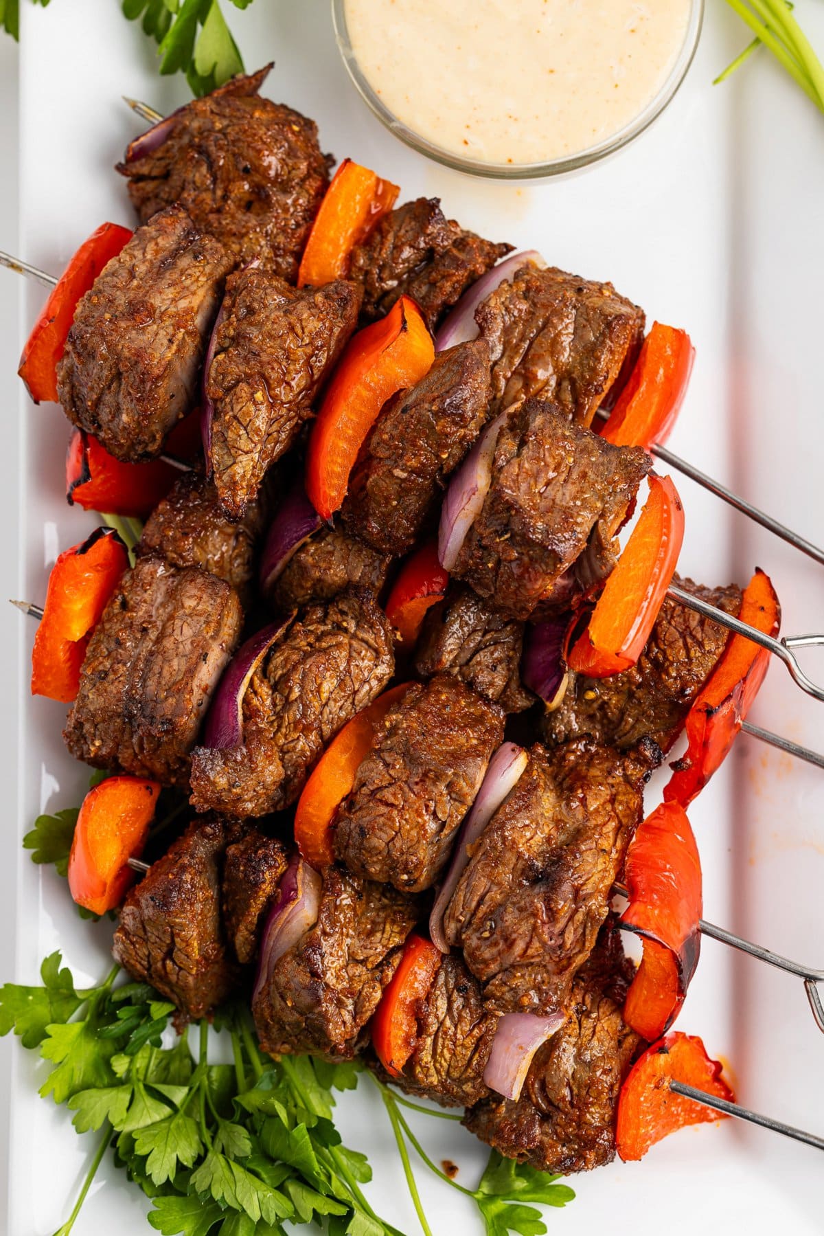 Broiled steak skewers on a platter with fresh parsley and creamy tahini sauce.