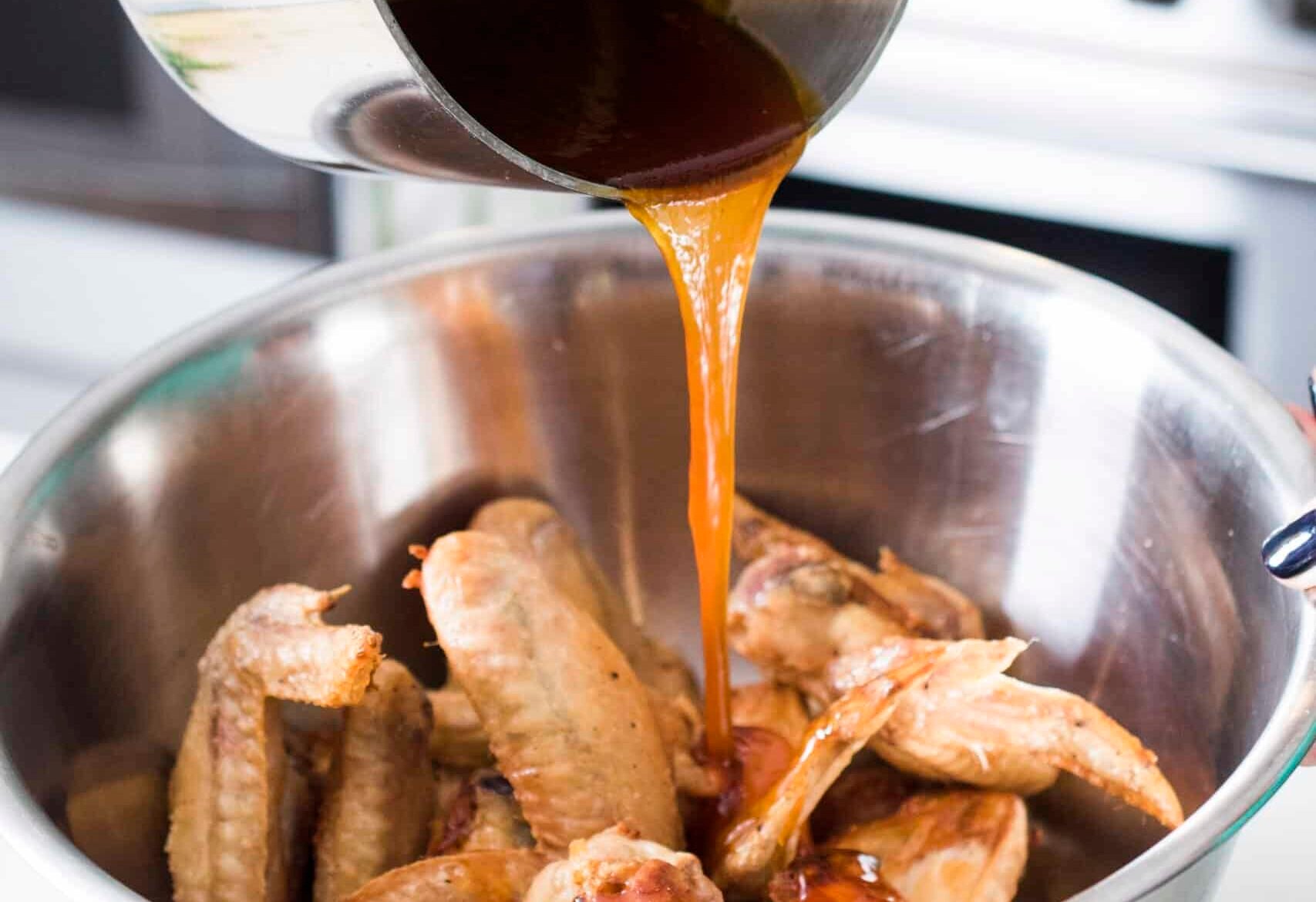 Pouring wing sauce over the top of cooked wings in a large metal bowl.