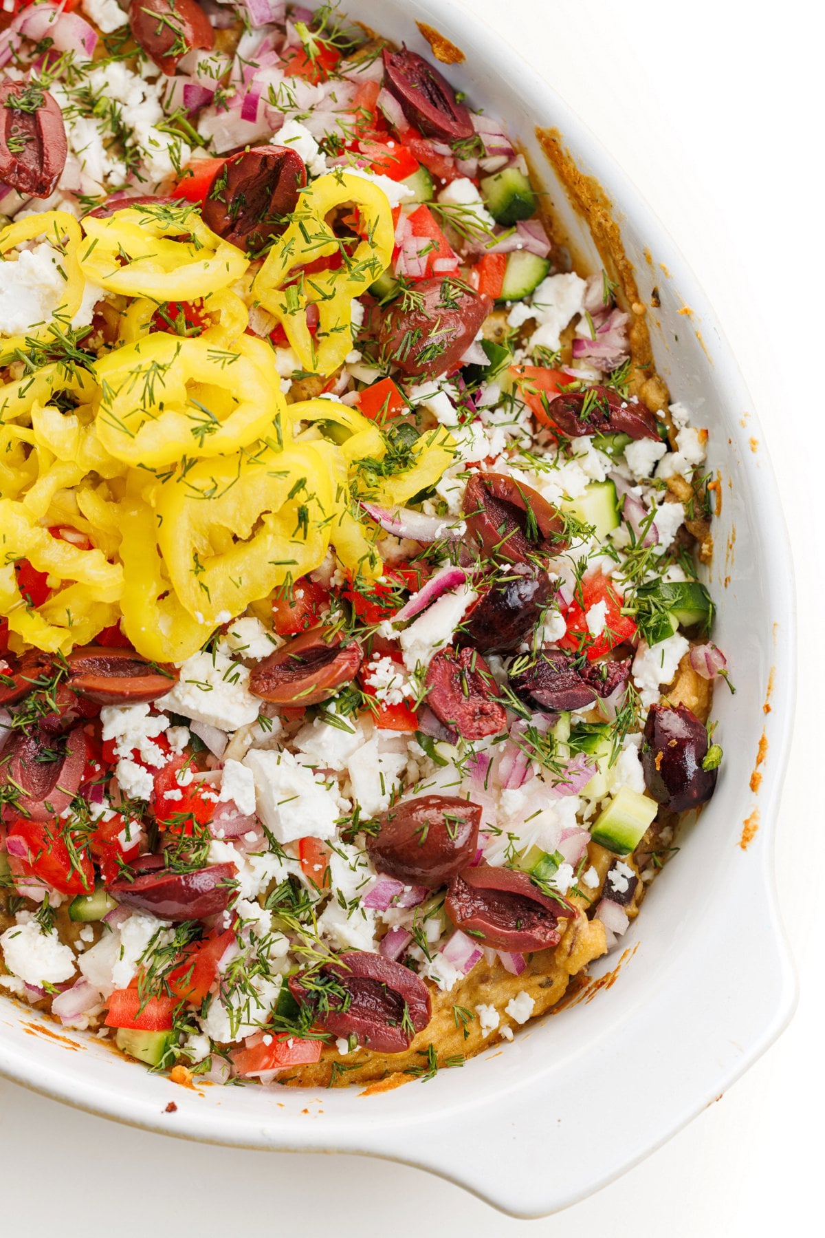 Greek baked hummus dip in dish topped with freshly chopped veggies and fresh herbs.