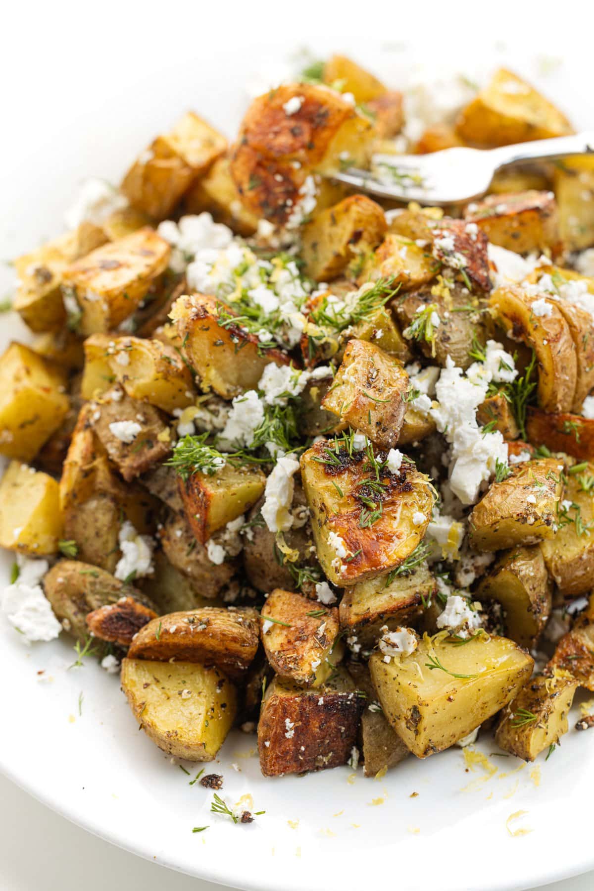 A fork through a Greek roasted potato on a plate full of roasted potatoes, garnished with feta cheese and herbs.