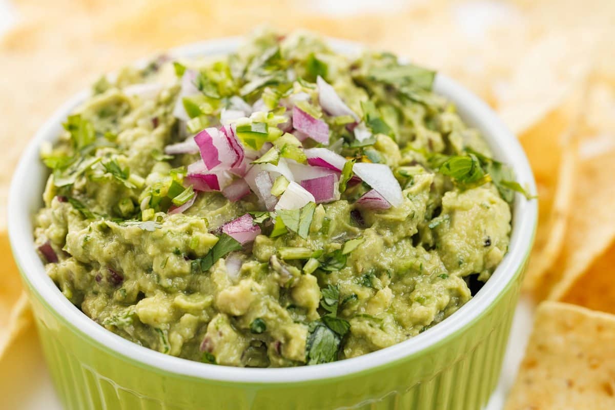Guacamole in green bowl with tortilla chips in background.