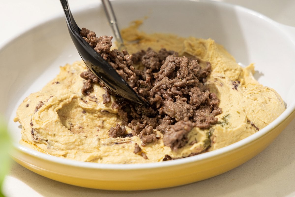 Adding cooked ground lamb to hummus in casserole dish.