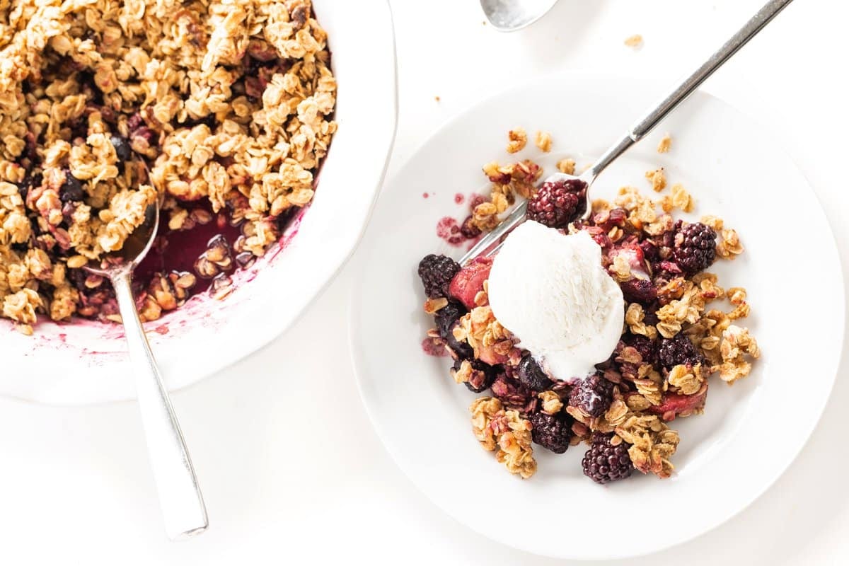 Pie dish of triple berry crisp with a scoop missing, the scoop is on a white plate next to it with a fork and ice cream.