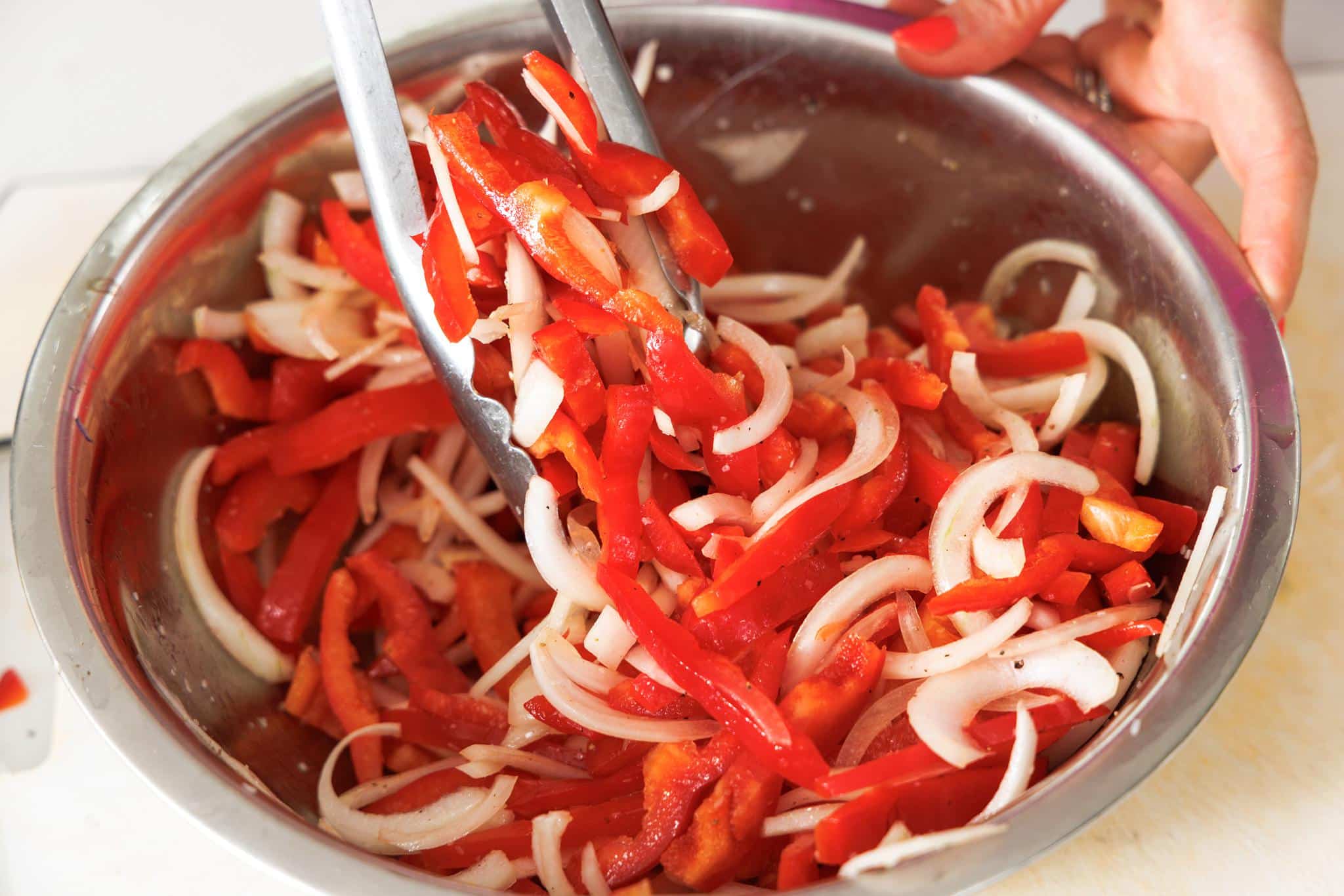 Tossing sliced bell peppers and onions in a bowl with tongs.