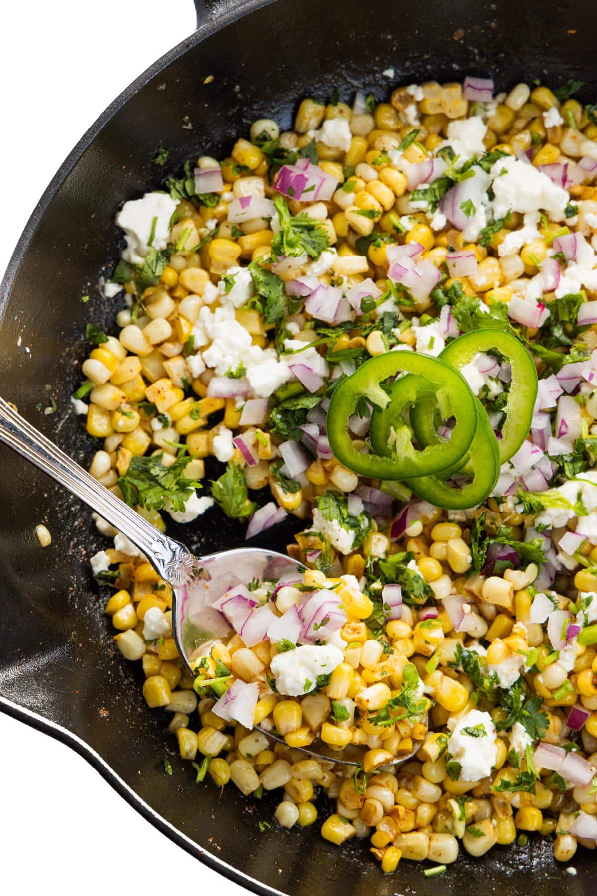 A spoon scooping out skillet Mexican street corn with red onion and cotija cheese.